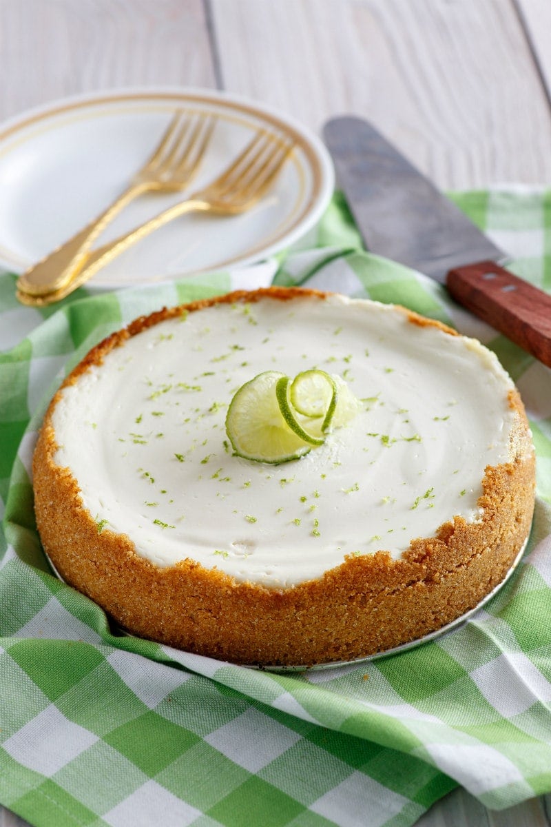 Key Lime Cheesecake sitting on a green and white checked tablecloth with serving plates, forks and serving spatula in the background