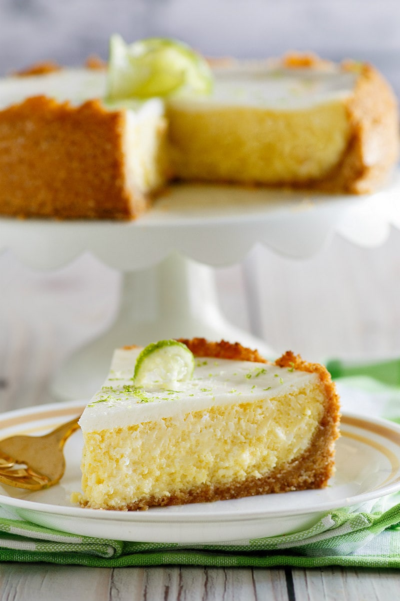 Slice of Key Lime Cheesecake on a white plate with a fork, set on a white and green napkin with cut cheesecake on display platter in the background