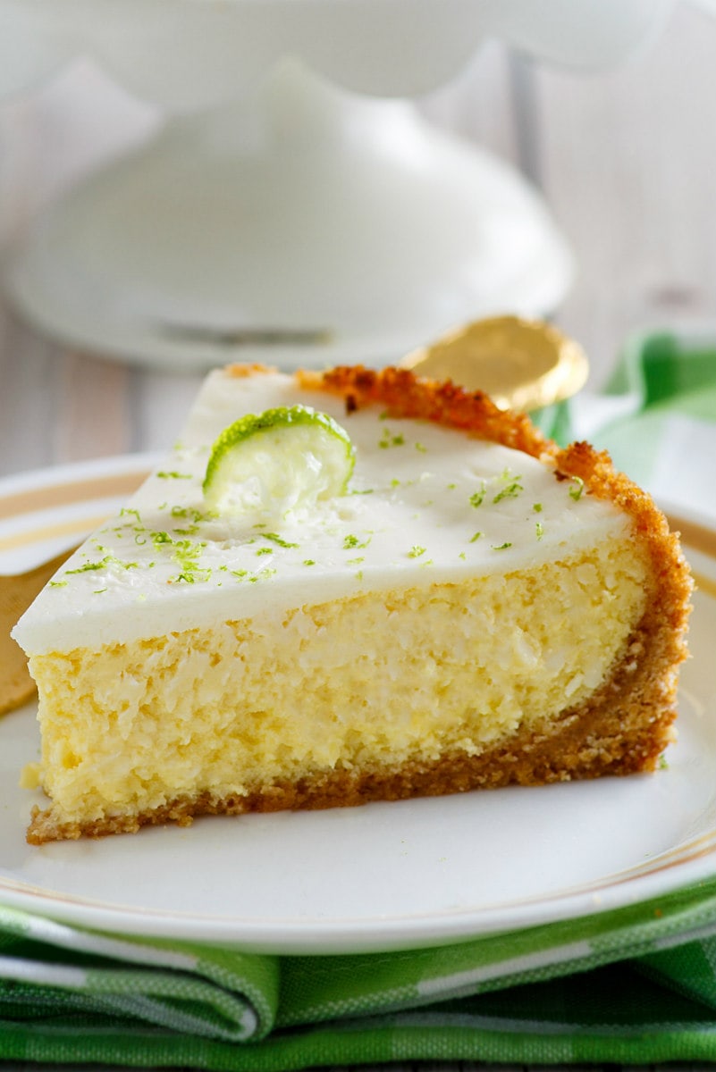 Slice of Key Lime Cheesecake on a white plate set on a green/white cloth with white display platter in the background
