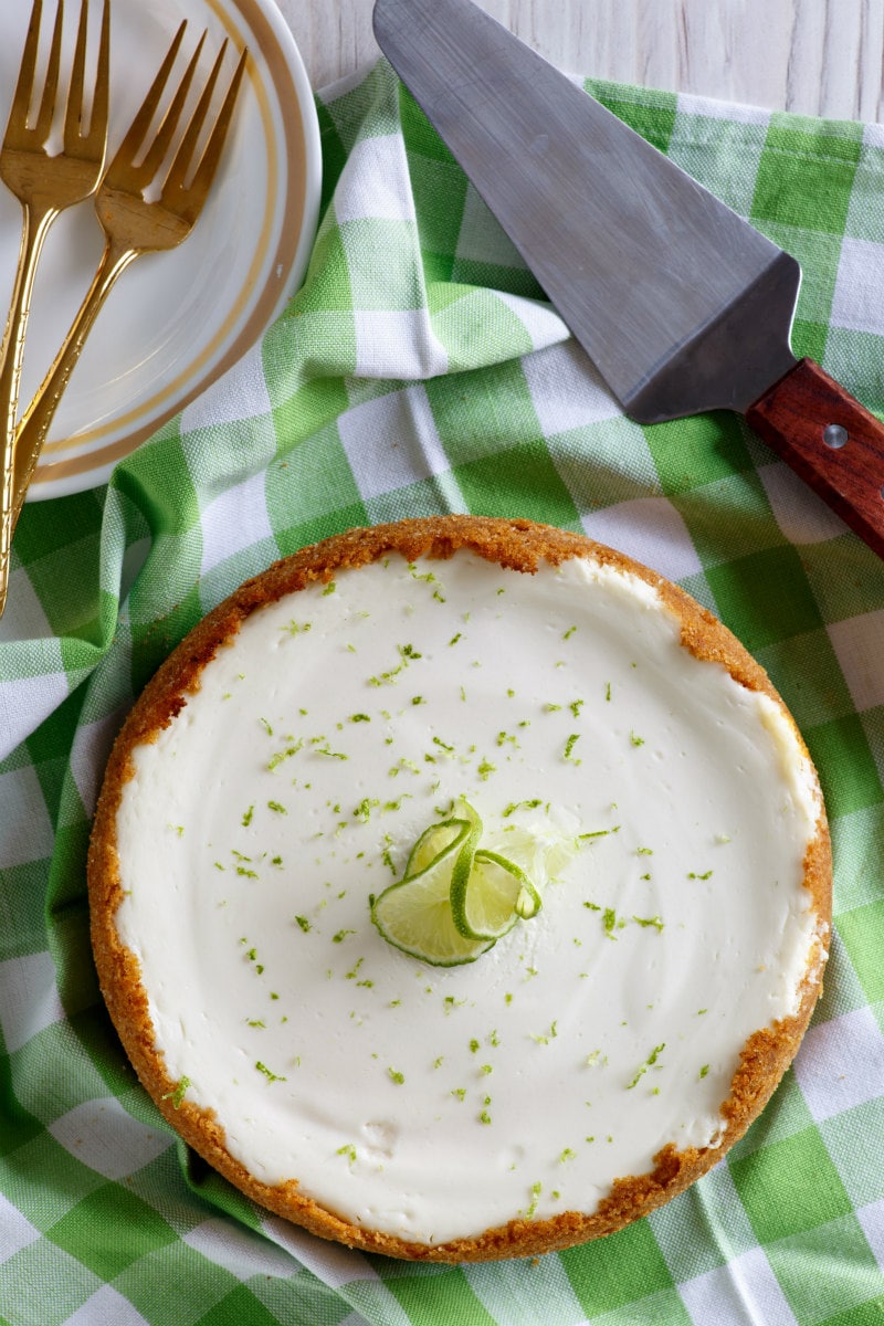 Key Lime Cheesecake sitting on a green and white checked tablecloth with serving plates, forks and serving spatula in the background