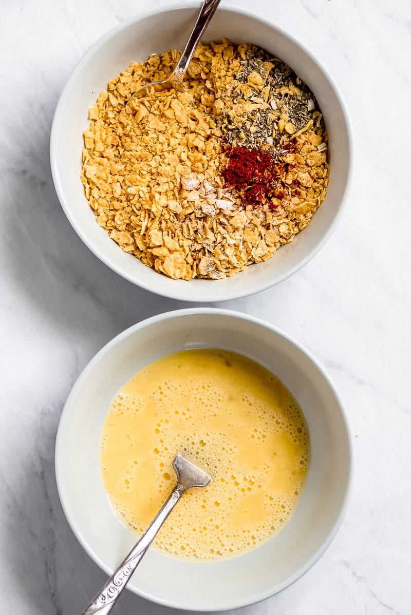 Overhead shot of ingredients for Cornflake Chicken- white dipping bowls of egg with a fork and cornflake crumbs plus spices with a spoon.