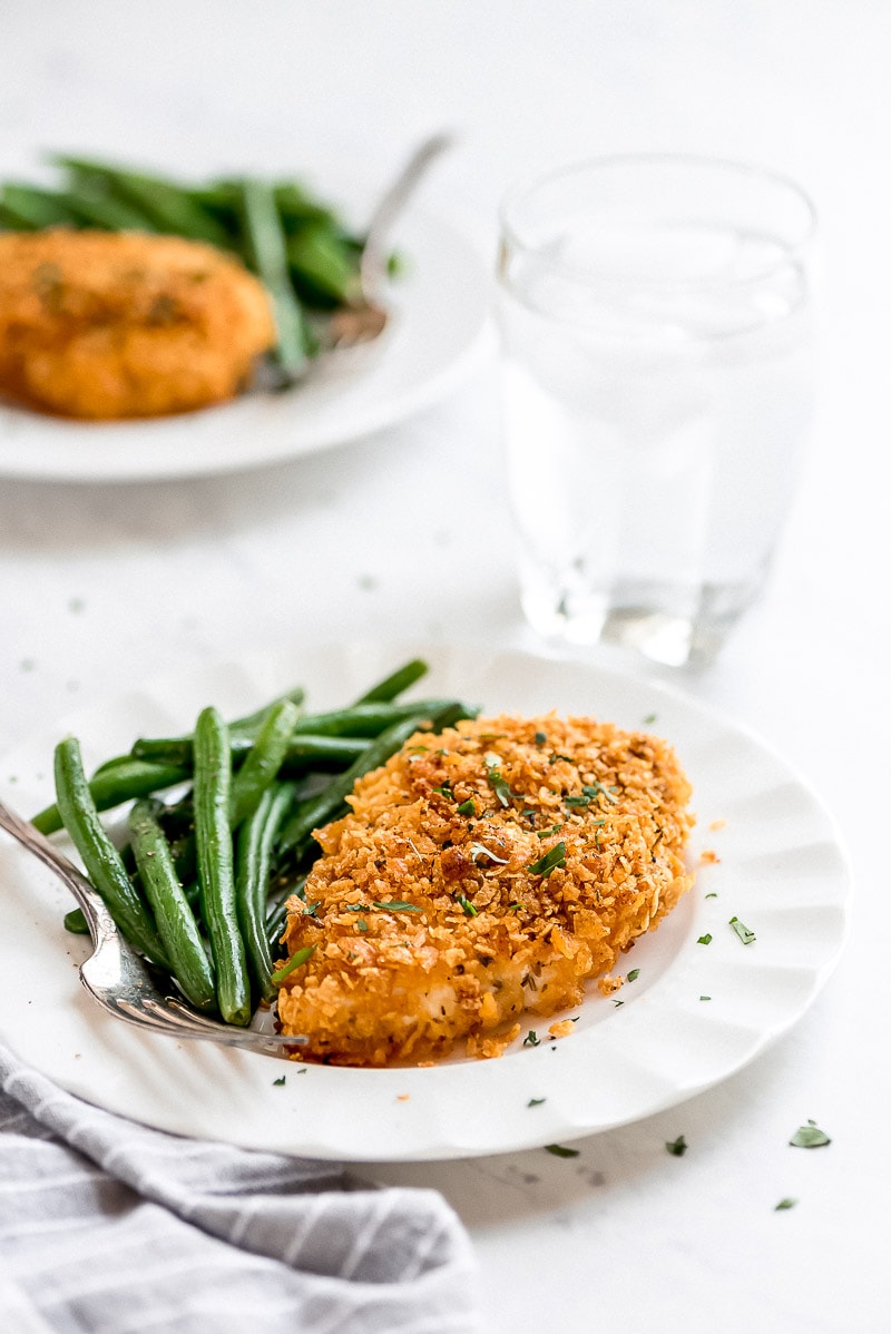 Baked Cornflake Chicken plates with green beans on a white plate. a second plate of cornflake chicken in the background. gray napkin and a glass of water also in view