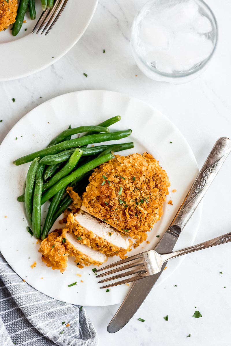 Baked Cornflake Chicken cut into slices on a white plate with green beans and a knife and fork.