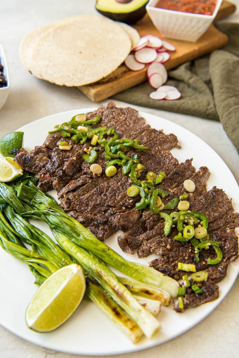 sliced carne asada on a white serving platter topped with jalapeno and with green onions, limes and corn tortillas on the side