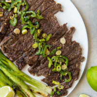 sliced carne asada on a white serving platter topped with jalapeno and with green onions and limes on the side