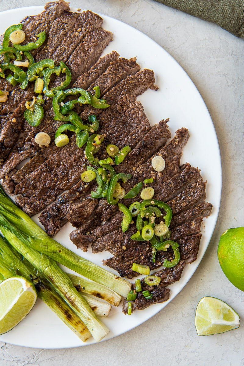 sliced carne asada on a white serving platter topped with jalapeno and with green onions and limes on the side
