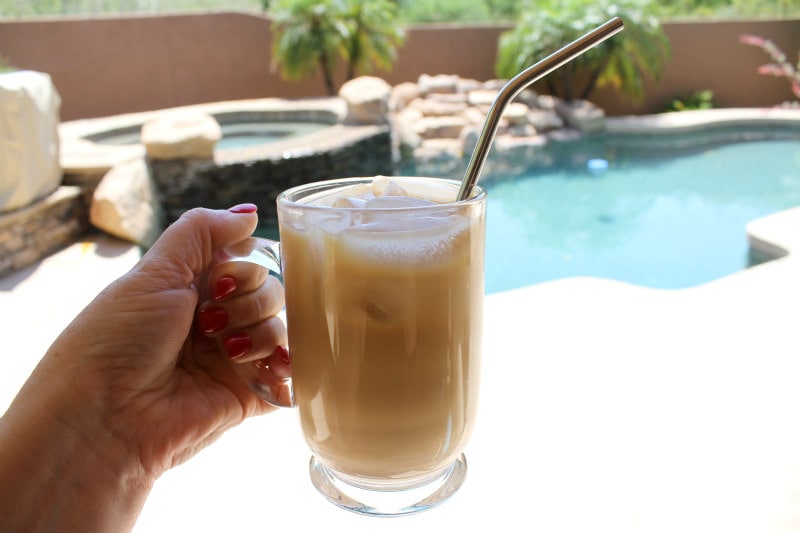 hand holding a mug of glass mug of cold brew coffee with a backyard pool scene behind it