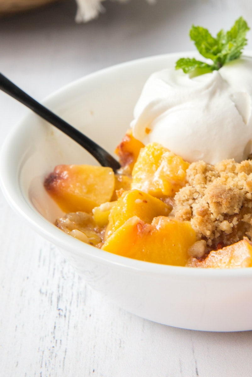 close up of a serving of peach cobbler french toast casserole in a white dish, garnished with whipped cream and fresh mint. Spoon in the bowl