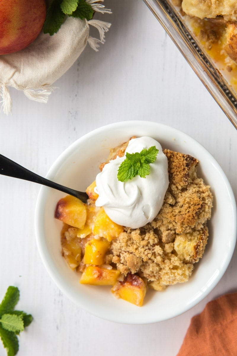 serving of peach cobbler french toast casserole in a white bowl garnished with whipped cream and fresh mint