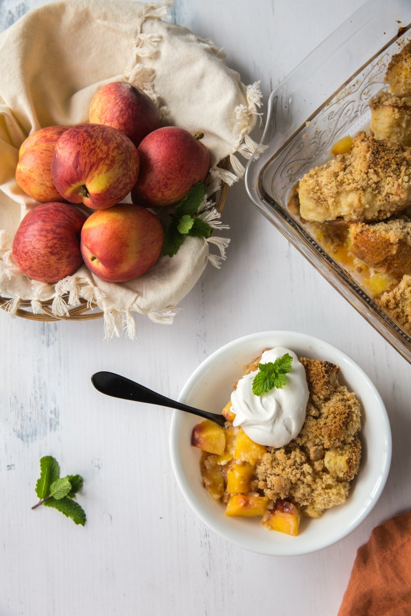 serving of peach cobbler french toast casserole in a white bowl garnished with whipped cream and fresh mint with basket of fresh peaches in the background