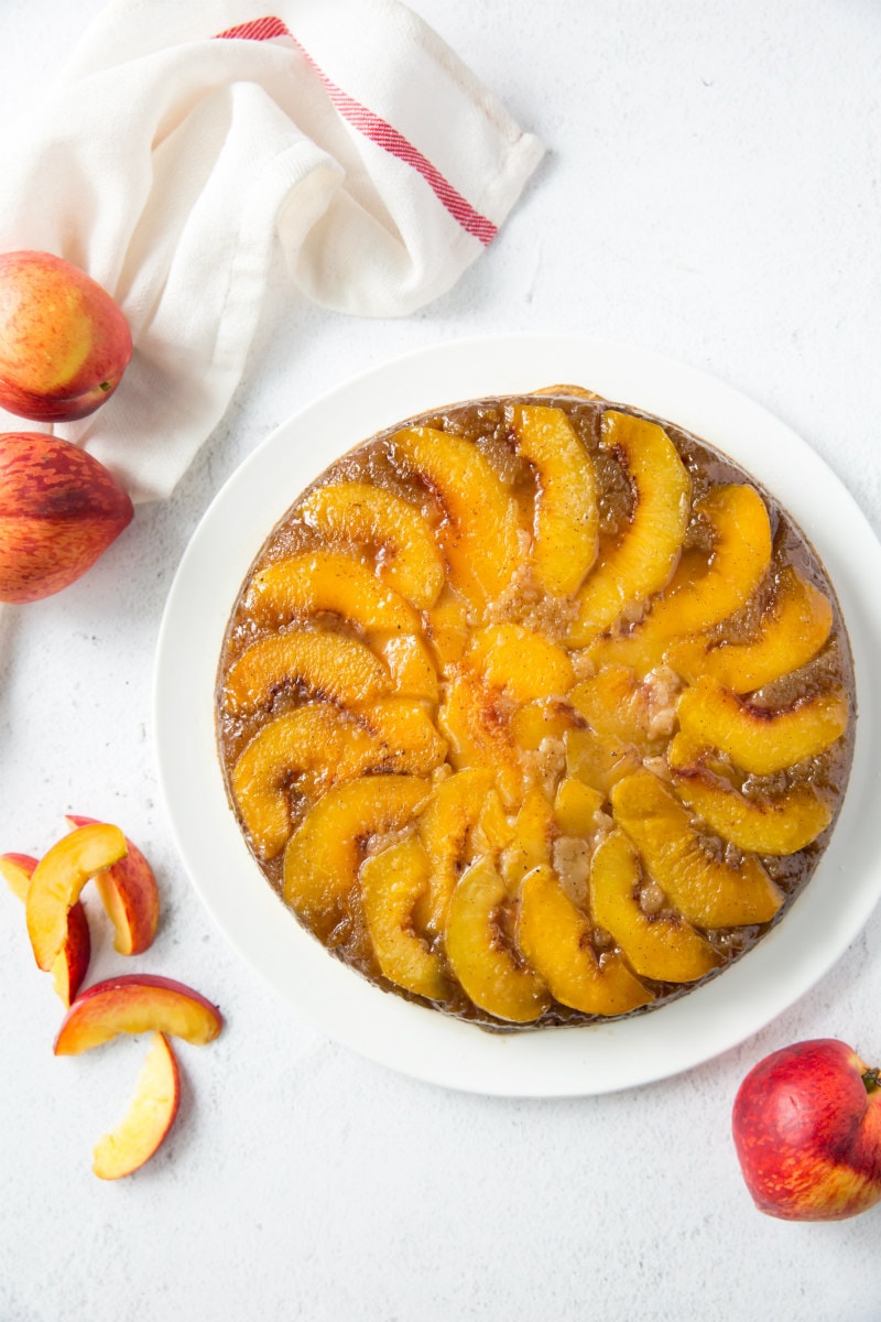overhead shot of peach upside down cake on a white plate with some fresh peaches in the background