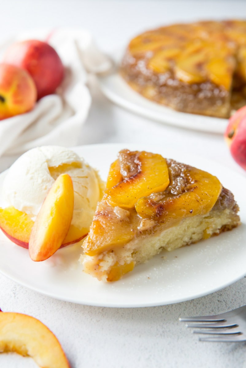 slice of peach upside down cake on a white plate with peaches and the cake in the background