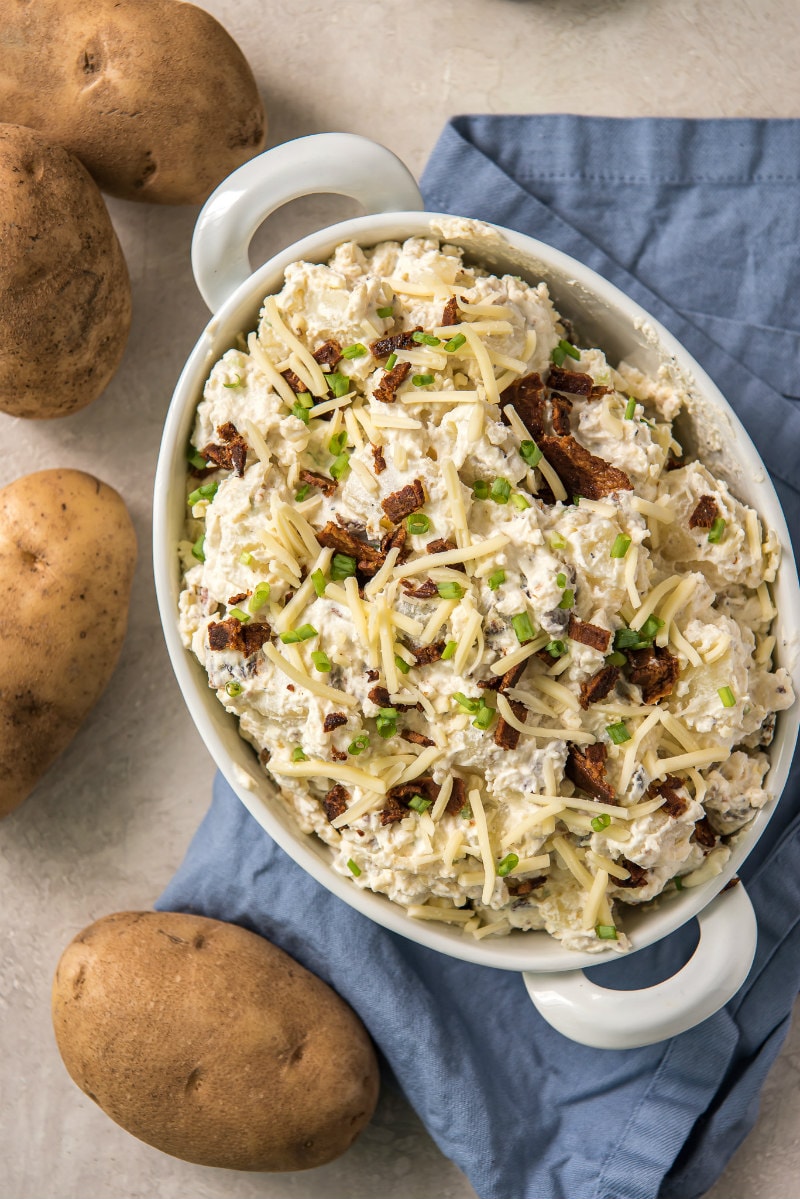 overhead shot of loaded baked potato salad in a white dish on a blue napkin. Potatoes in the background