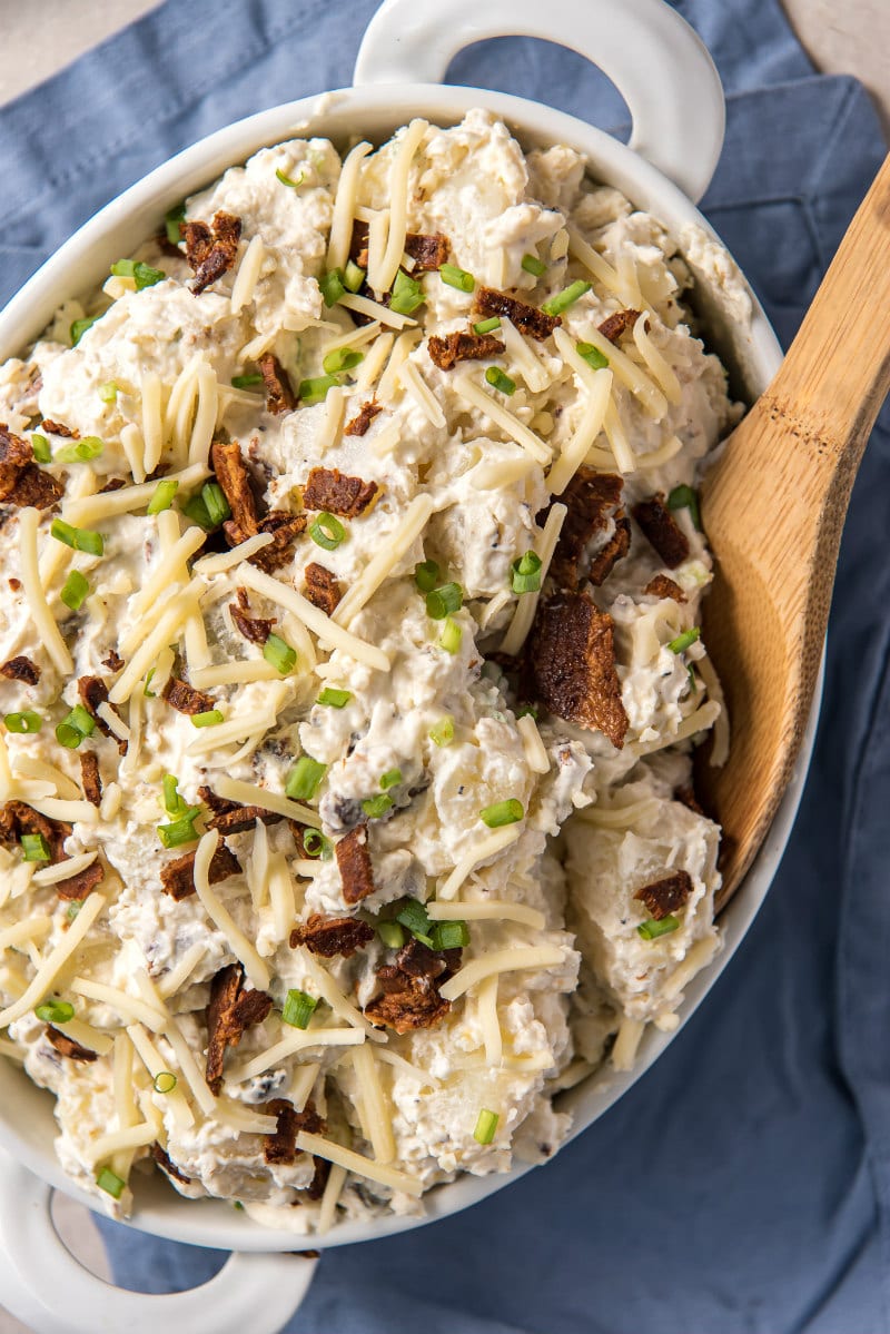 loaded baked potato salad in a white dish with a wooden spoon- on a blue napkin