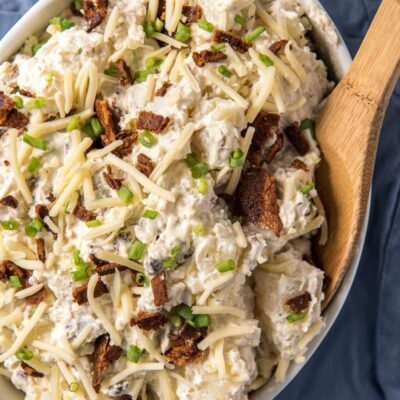 Loaded Baked Potato Salad in a white dish with a wooden spoon on a blue napkin