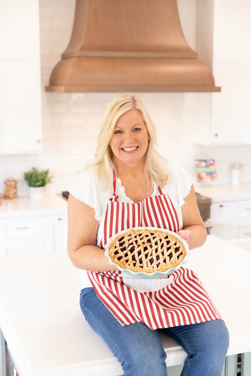 RecipeGirl sitting on the kitchen island wearing a red and white striped apron and holding a cherry pie