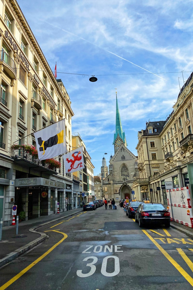 streets of Zurich, Switzerland: city street with cars parked on side and hotel with flags displayed. Church spier in background.