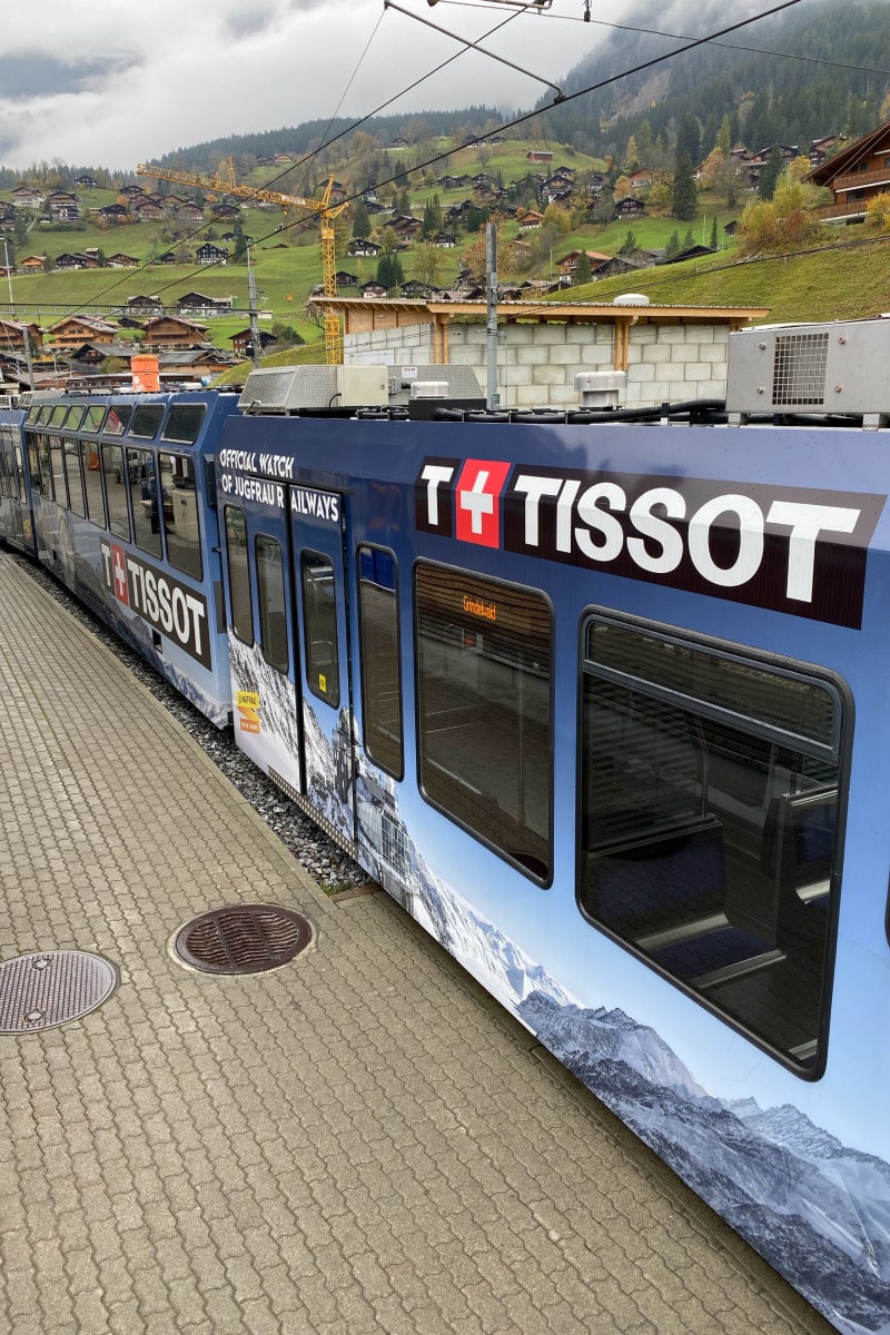 Blue Tissot Train to the Swiss Alps sitting at station. Green hills in the background.