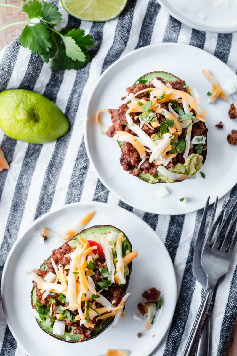 two avocado halves stuffed with taco filling on white plates. on a striped napkin with forks.