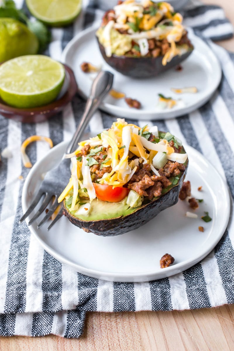 half an avocado stuffed with taco filling, sitting on a white plate with a fork. Lime in the background. Sitting on a blue and white striped napkin.