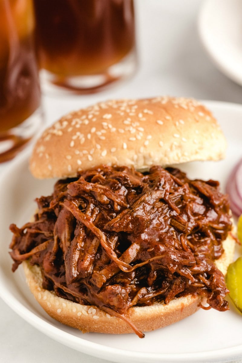 barbecue beef sandwich on a sesame seed bun displayed on a white plate with glasses of soda in the background