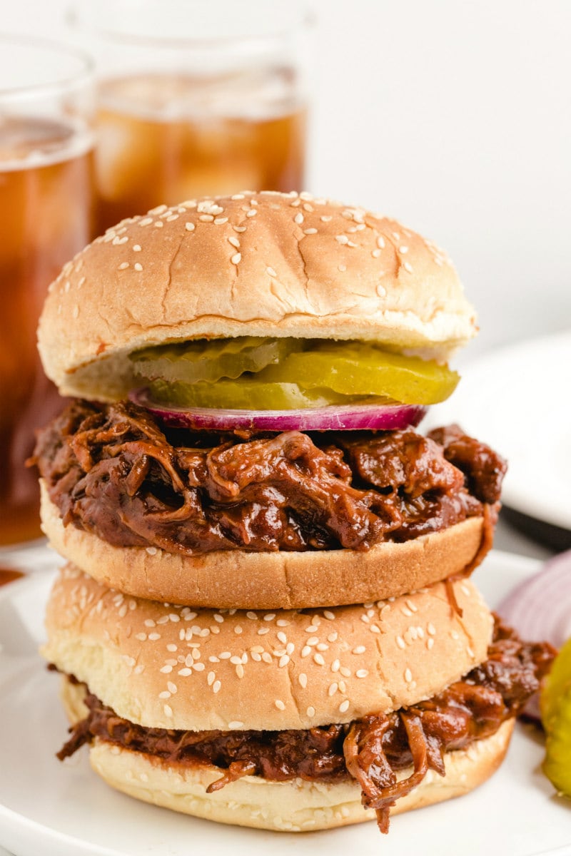 two barbecue beef sandwiches stacked on top of each other displayed on a white plate with glasses of soda in the background
