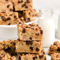 stack of three blondies with more blondies on a white platter in the background and on the sides. Glass of milk too.