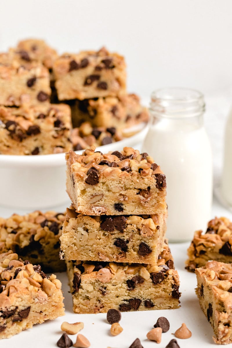 stack of three blondies with more blondies on a white platter in the background and on the sides. Glass of milk too.