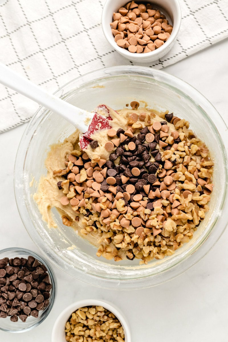 overhead shot of a glass bowl with blondie batter and a lot of chocolate chips. More chocolate chips and walnuts in bowls  on the side with a simple patterned napkin