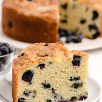 slice of buttermilk blueberry cake on a white plate with the rest of the cake on a white plate in the background