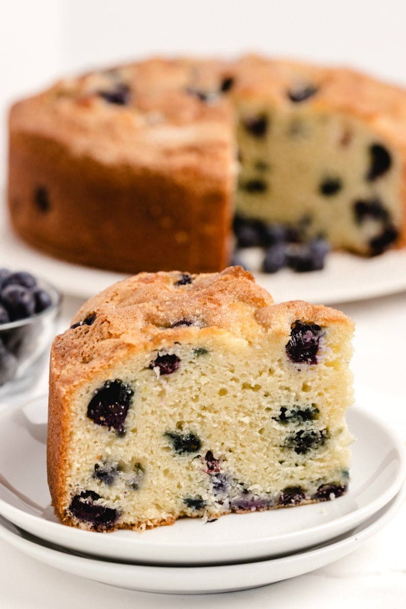slice of buttermilk blueberry cake on a white plate with the rest of the cake on a white plate in the background