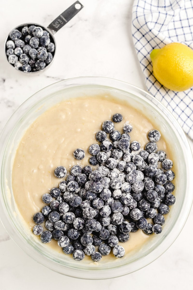 overhead shot of bowl of cake batter with blueberries added on top. more blueberries in a measuring cup on the side. lemon and gray/white dishtowel on the side too.