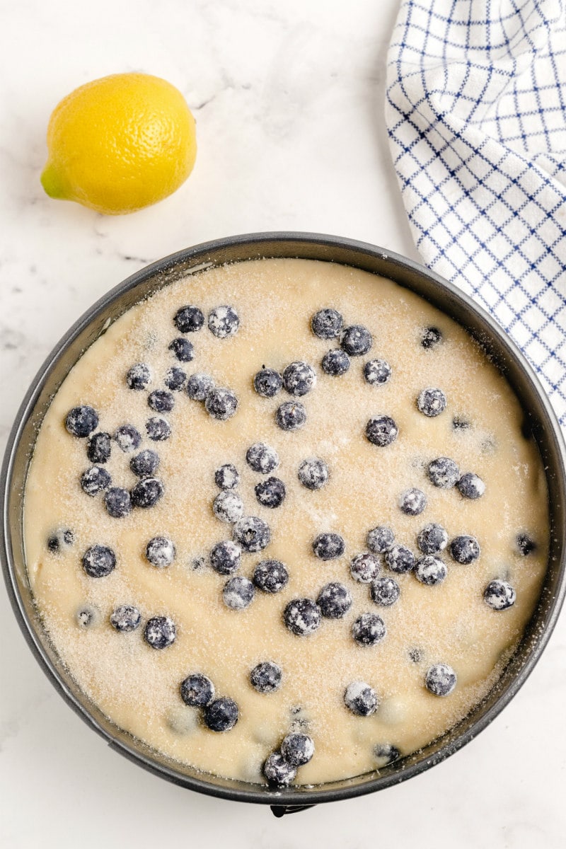 overhead shot of blueberry cake batter in a round pan with a white/gray plaid dishtowel and lemon in the background