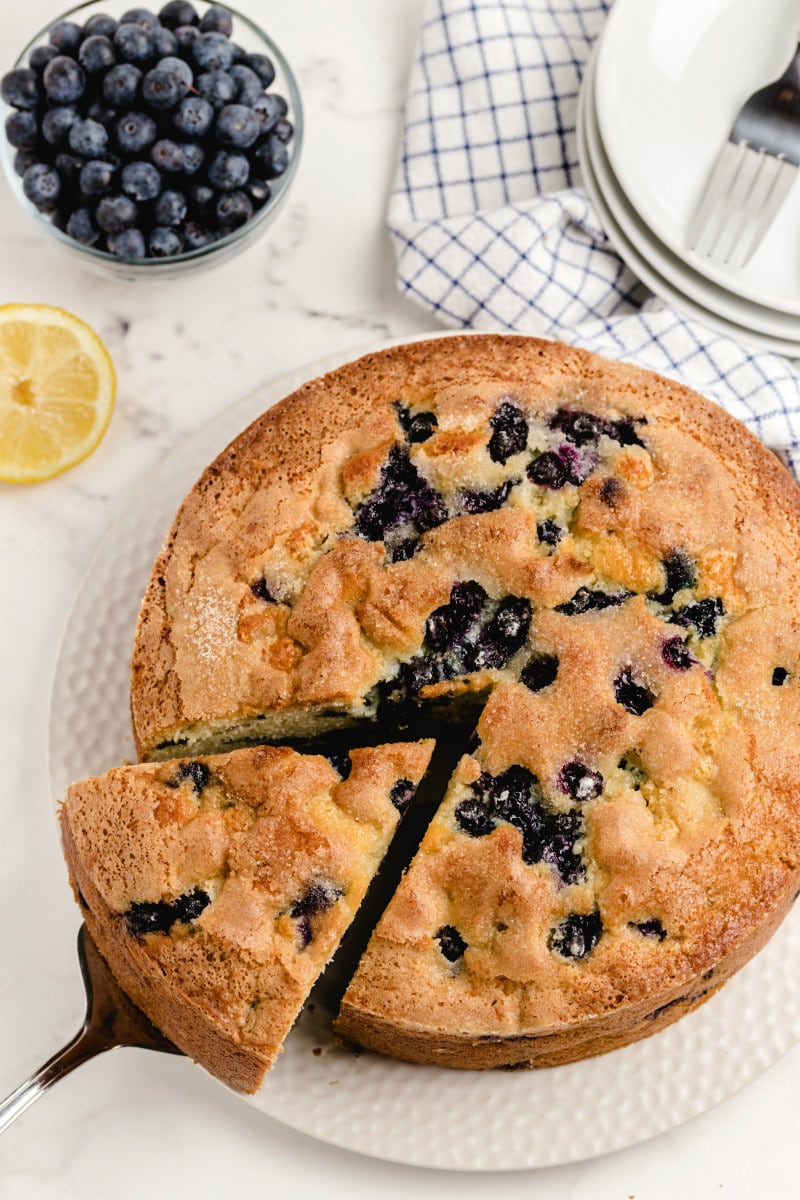 overhead shot of slice being taken out of buttermilk blueberry cake on a white plate. serving plates and forks sitting in the background on a gray and white plaid towel. half a lemon and bowl of fresh blueberries in the background.
