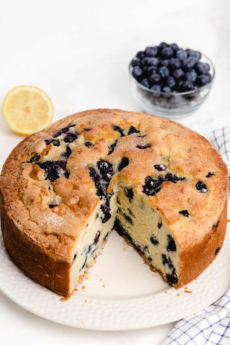 buttermilk blueberry cake with a big slice taken out of it on a white plate. half a lemon and bowl of fresh blueberries in the background