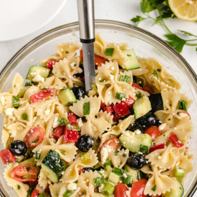 overhead shot of bow tie pasta salad in a glass bowl with a serving spoon and a half lemon and fresh herbs on the side