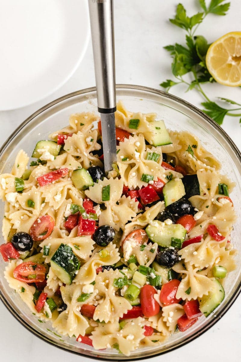 overhead shot of bow tie pasta salad in a glass bowl with a serving spoon and a half lemon and fresh herbs on the side