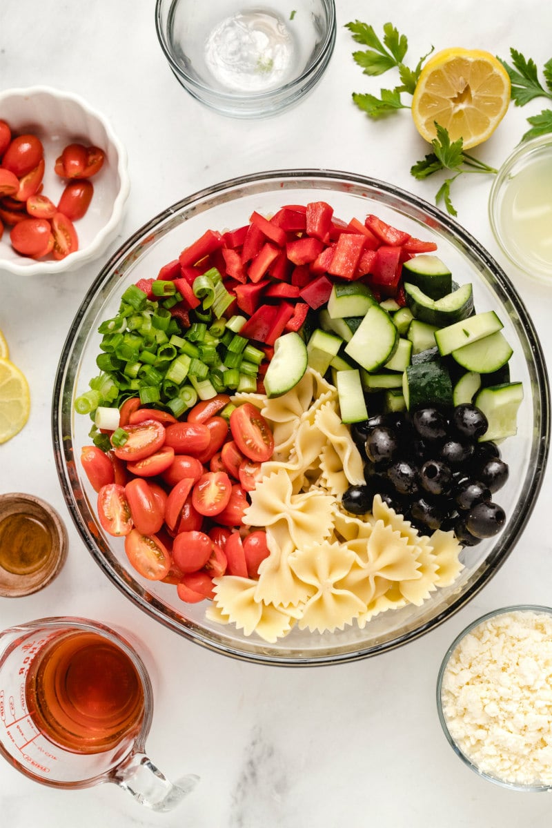 overhead shot of bow tie pasta salad ingredients in a glass bowl with dressing, cheese and tomatoes displayed in the background