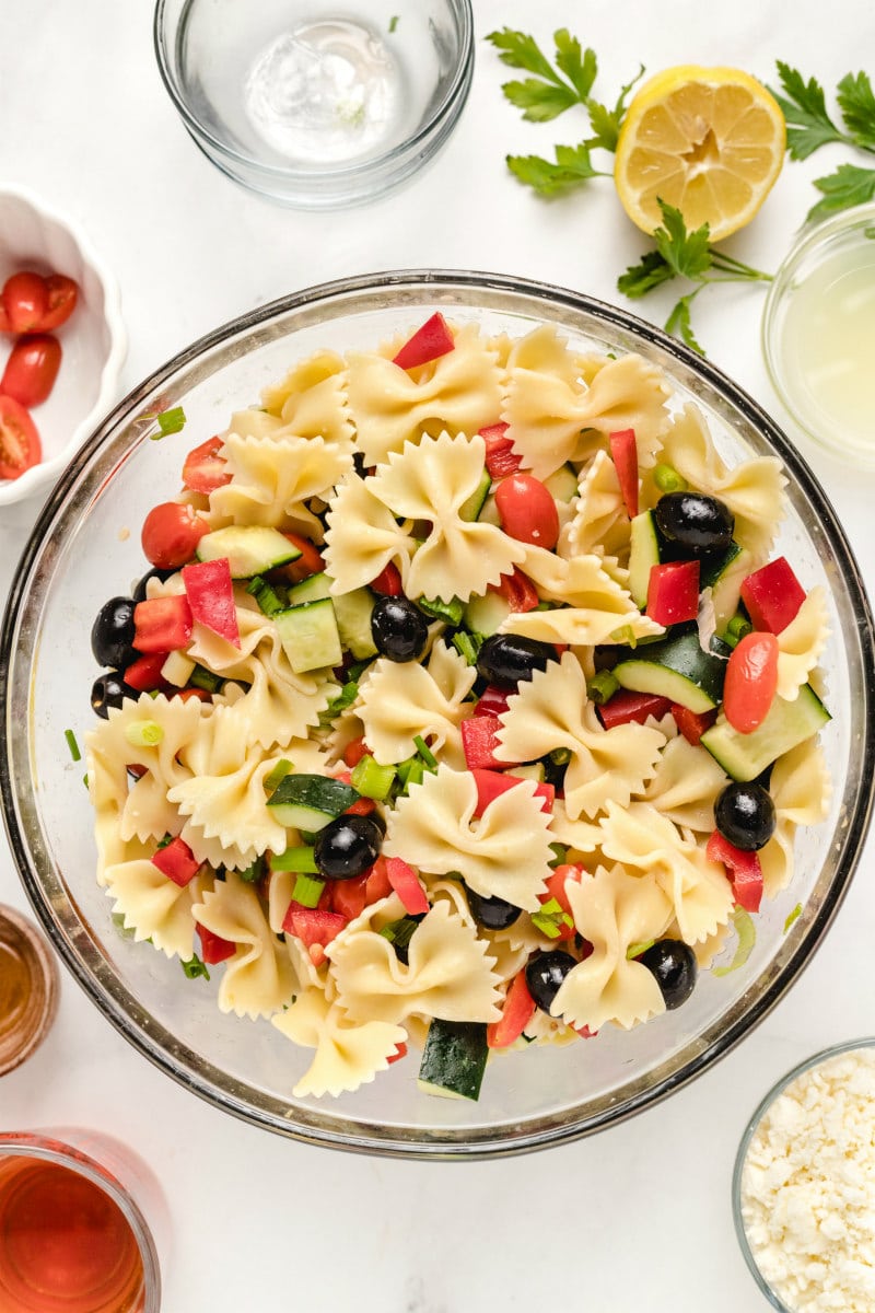 overhead shot of bow tie pasta salad in a glass bowl with bowls of tomatoes, cheese and dressing in the background