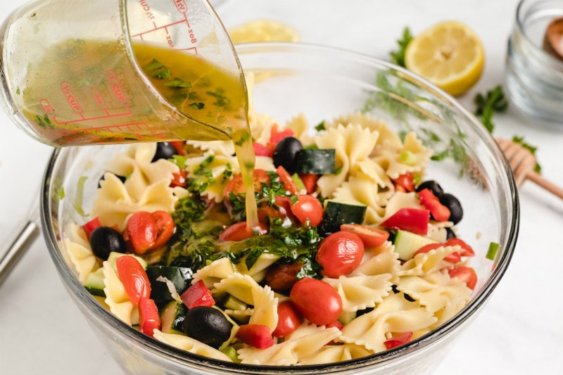 pouring vinaigrette from a measuring cup onto bow tie pasta salad in a glass bowl