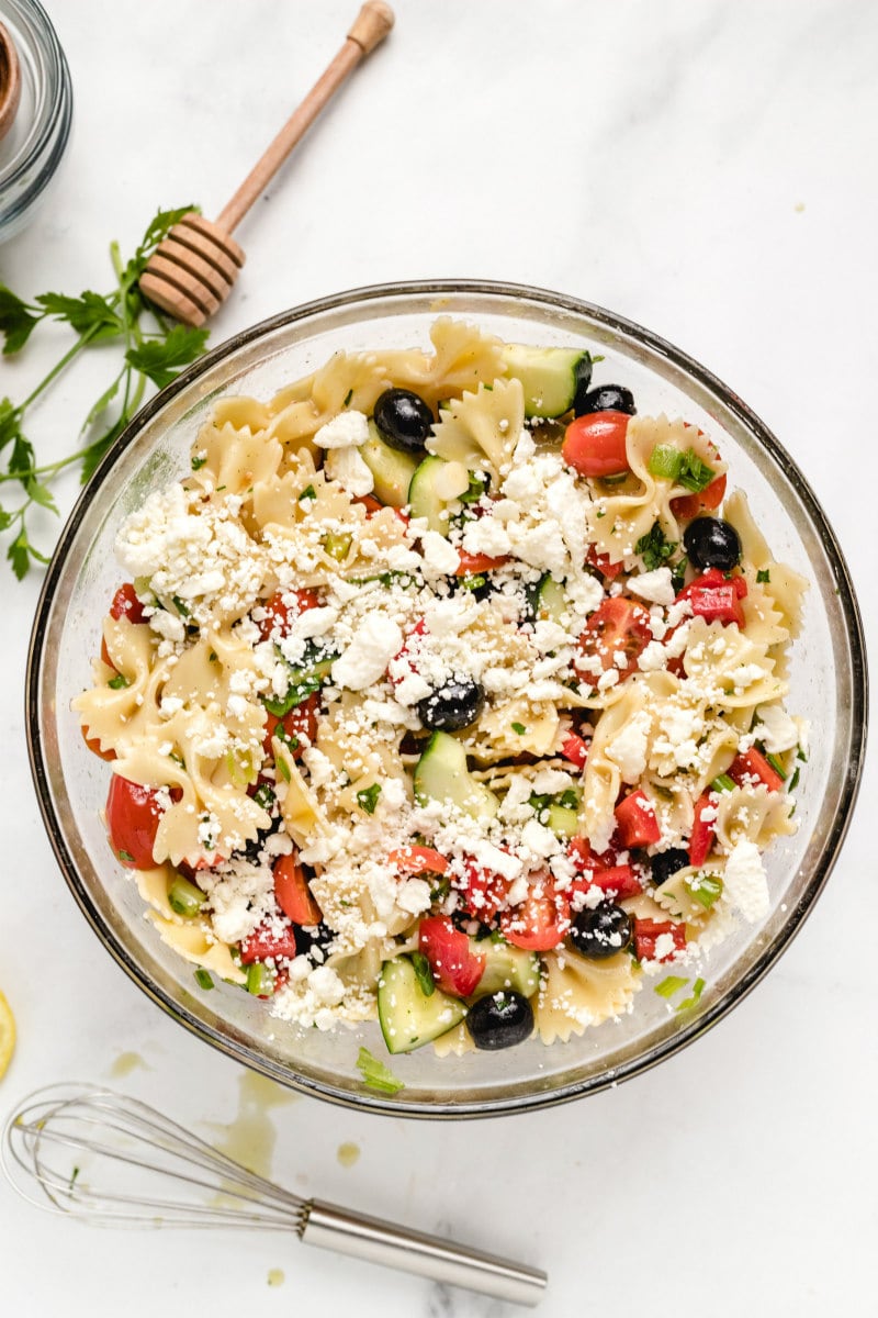 overhead shot of bow tie pasta salad in a glass bowl and feta cheese sprinkled on top. whisk and honey wand displayed in background