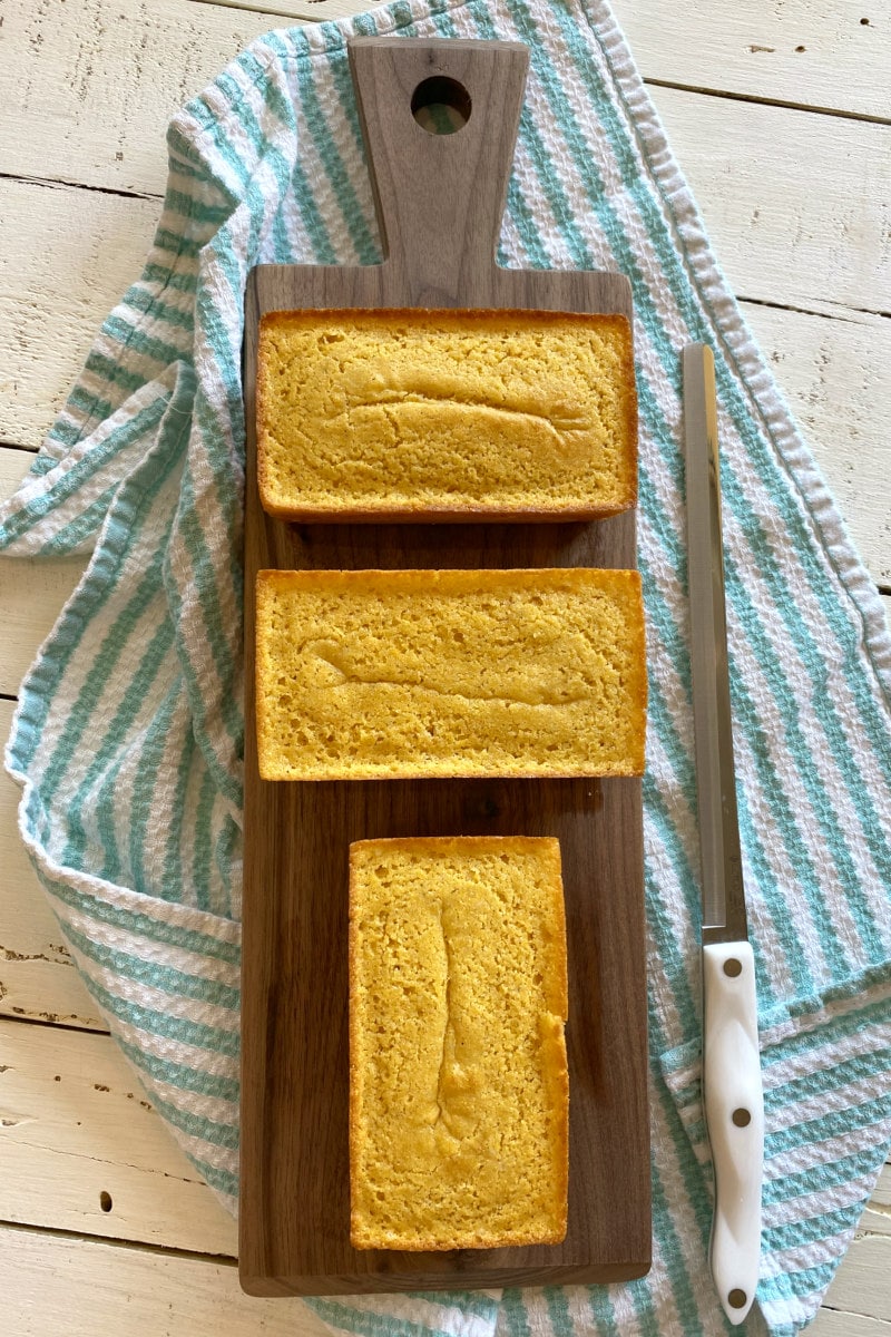 overhead shot of three mini loaves of buttermilk cornbread on a cutting board with a bread knife sitting alongside. set on a white/teal striped cloth napkin