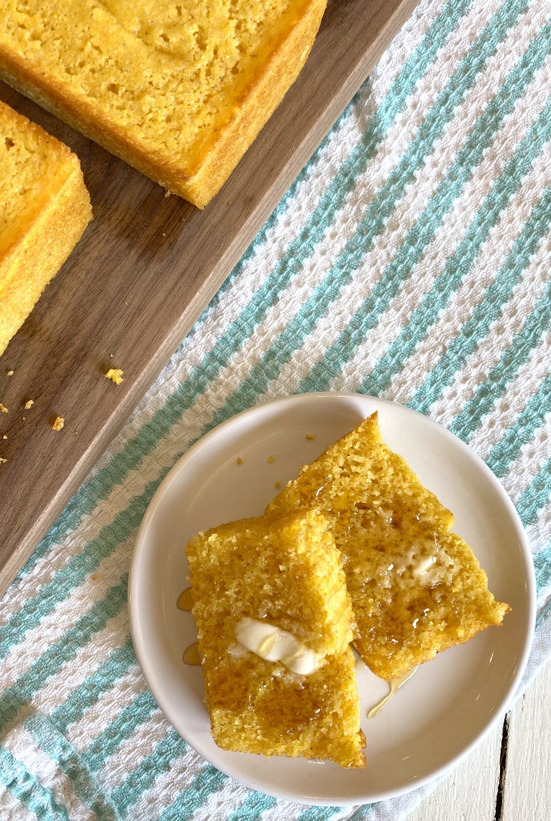 overhead shot of two slices of buttermilk cornbread on a white plate with butter and honey with a peek at more cornbread on a cutting board in the background- set on a white/teal striped cloth napkin