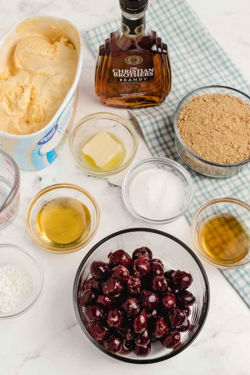 ingredients displayed for cherries jubilee ice cream pie in glass bowls: cherries, water, sugar, cornstarch, brandy, butter, honey, graham cracker crumbs and vanilla ice cream