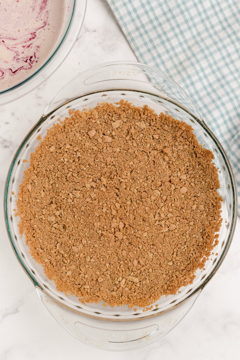 baked graham cracker crust in a pyrex pie plate with a peek at the ice cream filling for cherries jubilee ice cream pie in the background and a green/white plaid cloth napkin