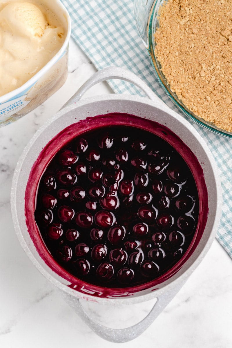 cherry sauce mixture for cherries jubilee ice cream pie in a white bowl. tub of vanilla ice cream and graham cracker crust in the background