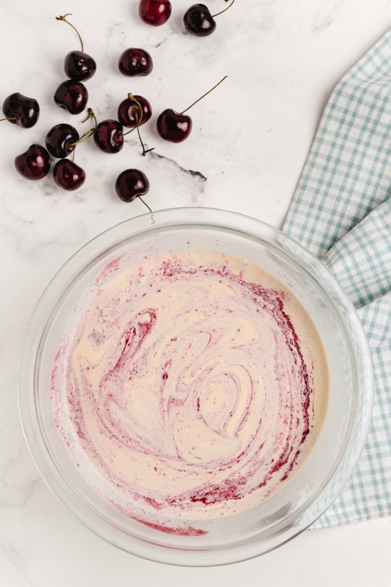 ice cream mixture for cherries jubilee ice cream pie in a glass bowl with a few fresh cherries scattered in the background and a green/white plaid cloth napkin