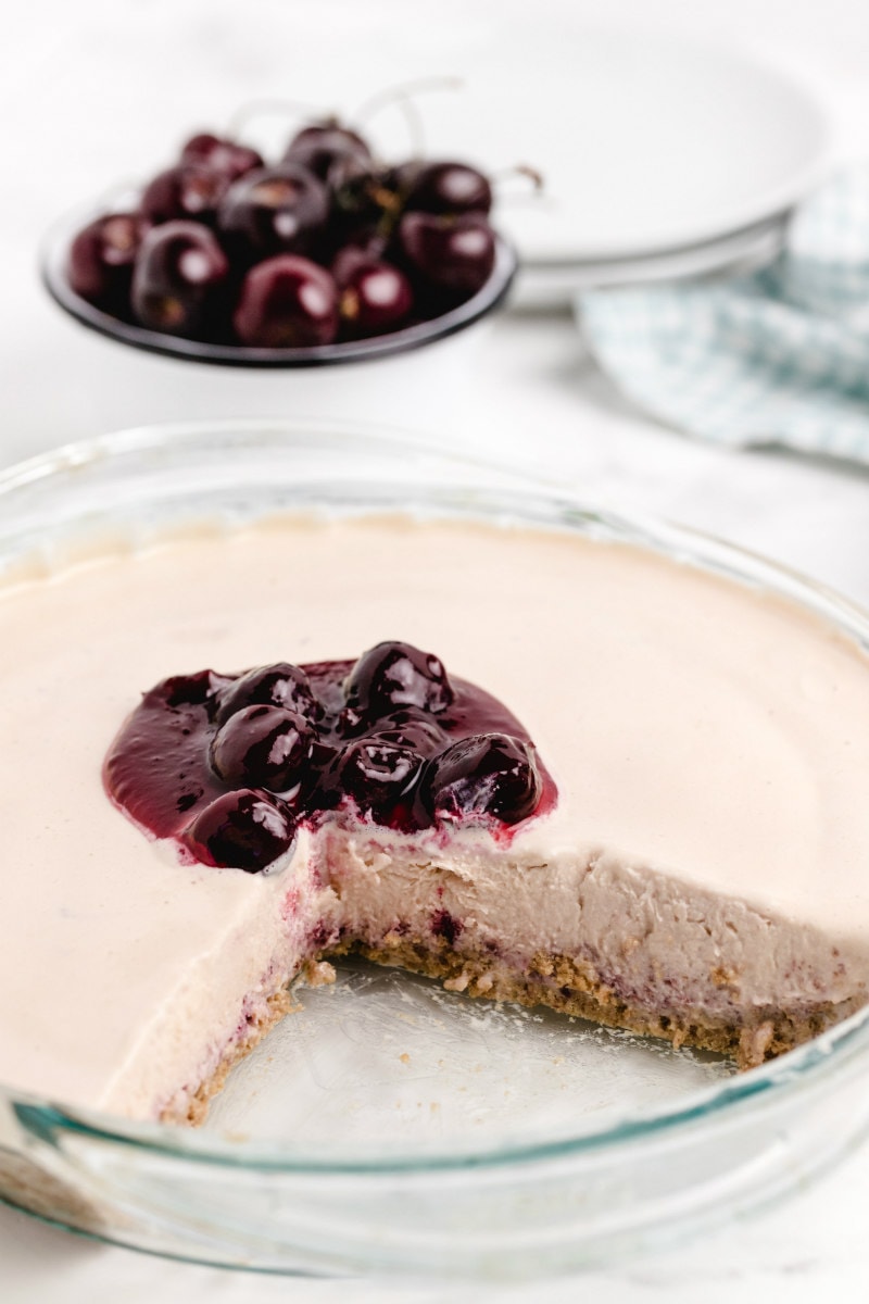 cherries jubilee ice cream pie in a pyrex pie plate with a slice cut out of it, cherry sauce on top and a bowl of fresh cherries in the background with stack of white plates and green/white checked cloth napkin