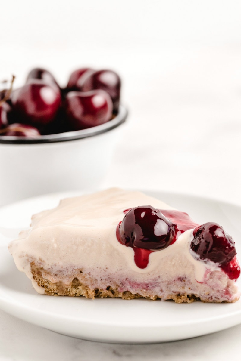 slice of cherries jubilee ice cream pie on a white plate with a bowl of fresh cherries in the background