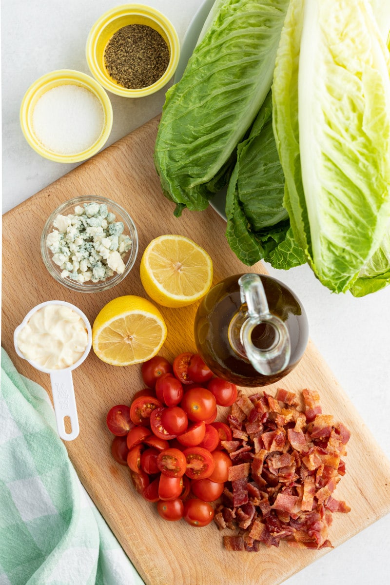 ingredients displayed for grilled romaine salad: romaine lettuce, mayonnaise, blue cheese, halved lemons, olive oil, tomatoes and bacon